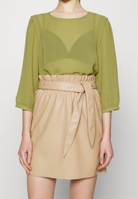 Grüne, transparente Bluse mit Puffärmeln über einem beigen Minirock aus Kunstleder, der eine taillierte Passform und eine glatte Textur aufweist.