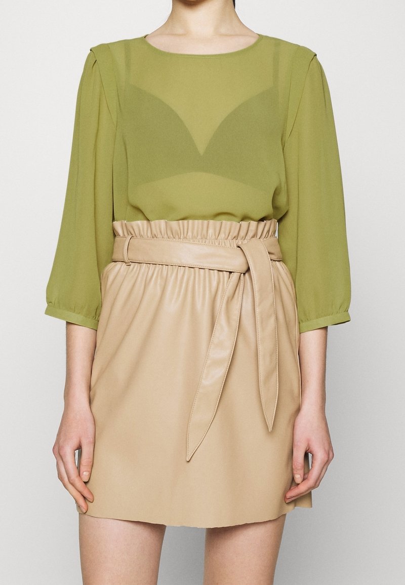 Grüne, transparente Bluse mit Puffärmeln über einem beigen Minirock aus Kunstleder, der eine taillierte Passform und eine glatte Textur aufweist.