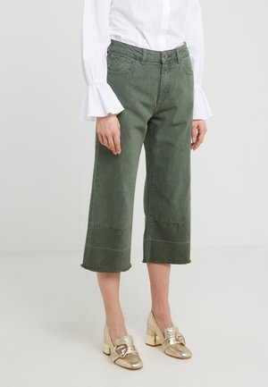Vrouw in olijfgroene, cropped jeans, witte blouse met uitlopende mouwen en metallic gouden blokhak schoenen met hartvormige gespen.