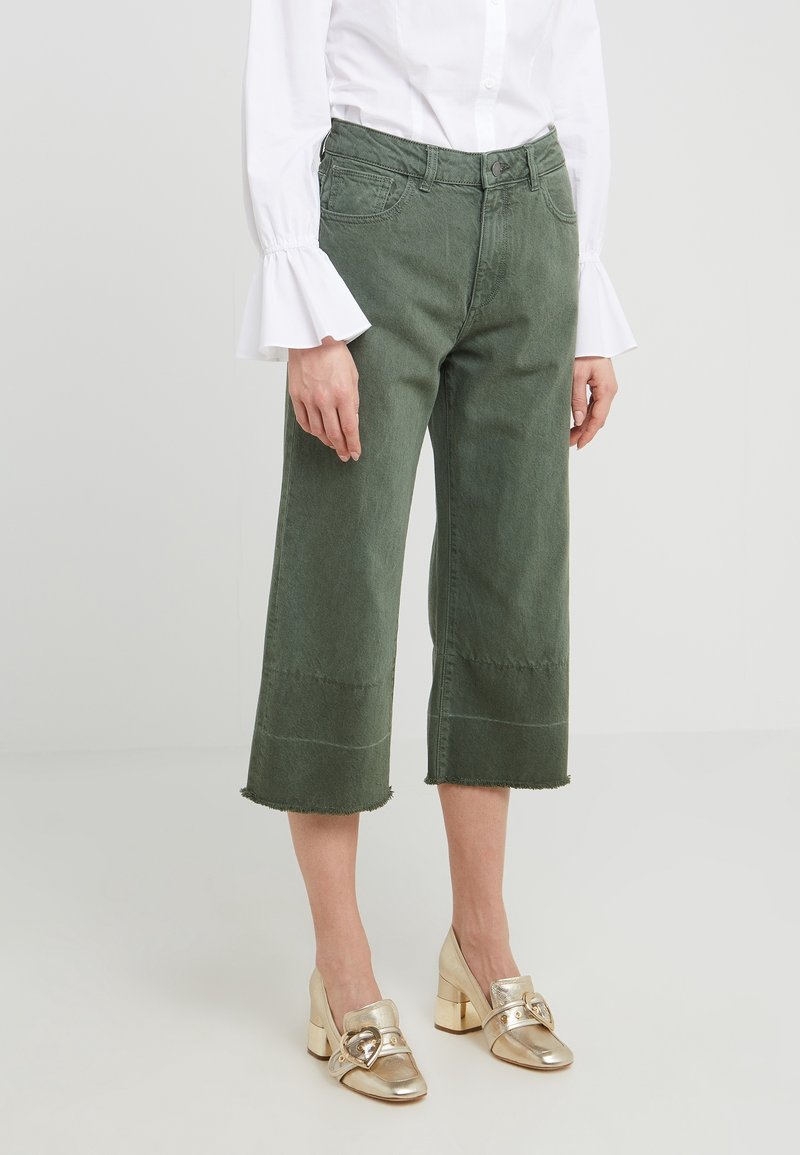 Nainen pukeutuneena vaaleanvihreisiin lyhyisiin farkkuihin, valkoiseen puseroon, jonka hihoissa on kellot, sekä metallinhohtoisiin kultaisiin paksupohjaisiin kenkiin, joissa on sydämen muotoiset soljet.