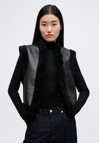 Μαύρο δερμάτινο γιλέκο με χνουδωτή μαύρη επένδυση και χωρίς μανίκια. Φορεμένο πάνω από μια εφαρμοστή μαύρη ζακέτα με ψηλό γιακά, συμπληρώνοντας με σκούρες τζιν παντελόνες.