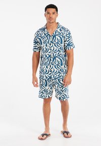 Conjunto de camisa de manga corta y pantalones cortos en blanco con un patrón de hojas azules. Tejido ligero, ajuste relajado, combinado con sandalias oscuras.