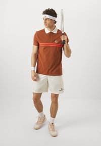 Rudas polo marškinėliai su balta apykakle ir raudonomis horizontaliomis juostomis, kartu su baltais šortais su logotipu ir rusvais sportbačiais. Laiko teniso raketę.