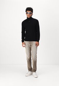 Črn pulover s turtleneck ovratnikom, svetle bež ožjene hlače in bele superge. Model nosi sončna očala sivih odtenkov s športnim dizajnom.