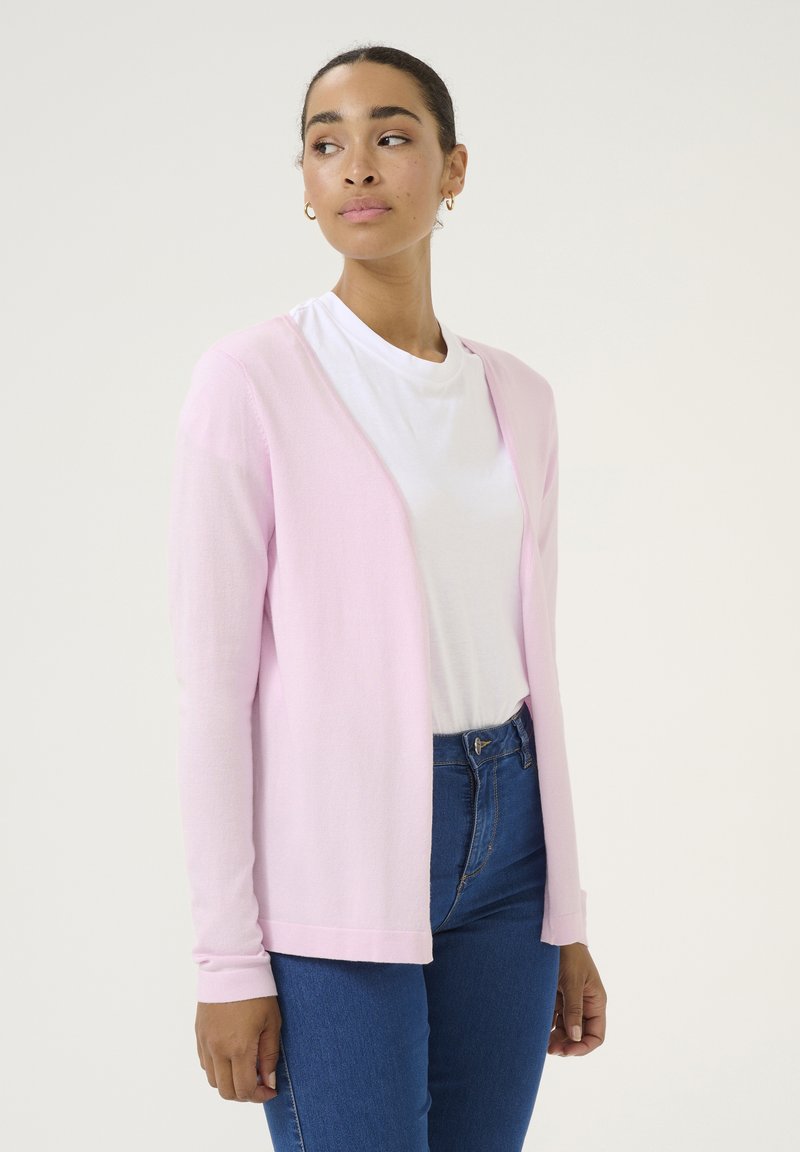 Cardigan rosa chiaro realizzato in morbido materiale knit, con apertura frontale, maniche lunghe e vestibilità comoda, abbinato a una maglietta bianca e jeans.