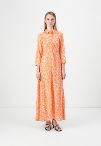Maxime-käytävä oranssista puuvillasta, jossa on valkoinen aaltoilu, edessä nappisulku ja korkea kaulus. Ominaisuuksina pitkät hihat ja virtaava siluetti.