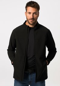 Sort softshell-jakke med høj krave, lynlåslomme på brystet og to sidelommer. Båret over en sort skjorte med en glat tekstur.