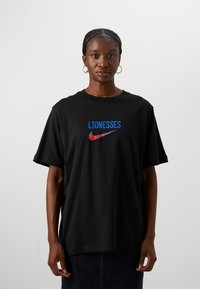 Camiseta de algodón negra con la palabra "LIONESSES" en azul y el logo de Nike en rojo. Corte oversized, con mangas cortas y cuello redondo.