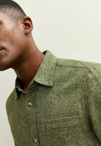 Chemise verte en tissu texturé, avec un devant à boutons et une poche poitrine. Elle présente un col pointu et des boutons marron clair.