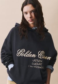 Námornícky mikina s bielym textom "Golden Essence" a "Urban Culture San Francisco", s prednou vreckom a vrstvenou svetlomodrou košeľou pod ňou.