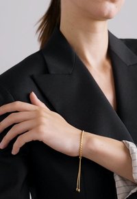 Gold twisted bracelet on a wrist, featuring two dangling beads. Worn with a black blazer over a light striped shirt.