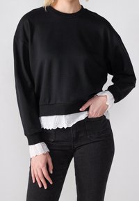 Sweat-shirt noir au design court, avec des manches longues et un col côtelé. En dessous, une chemise blanche avec un bord festonné en broderie anglaise est visible.