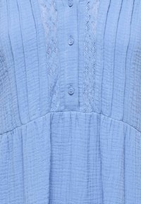 Material texturat albastru deschis cu panouri verticale din dantelă și nasturi mici, rotunzi, în partea din față a unei haine.