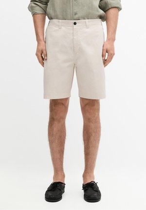 Mann trägt beige knielange Shorts, olivgrünes langärmliges Hemd mit hochgekrempelten Ärmeln und schwarze Schnürschuhe, stehend.