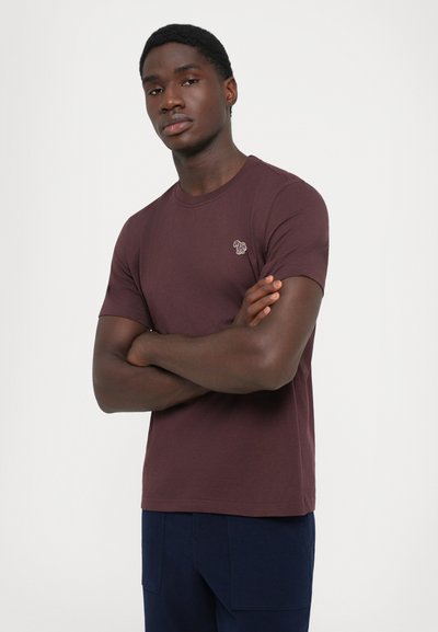 Jonge man met gekruiste armen, gekleed in een bordeauxrode t-shirt met korte mouwen en een klein geborduurd logo, en donkere blauwe broek.