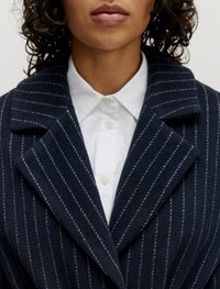 Marineblauwe wollen jas met witte streepjes, voorzien van een geknipte kraag en gestructureerde revers, gedragen over een witte overhemden met knopen.