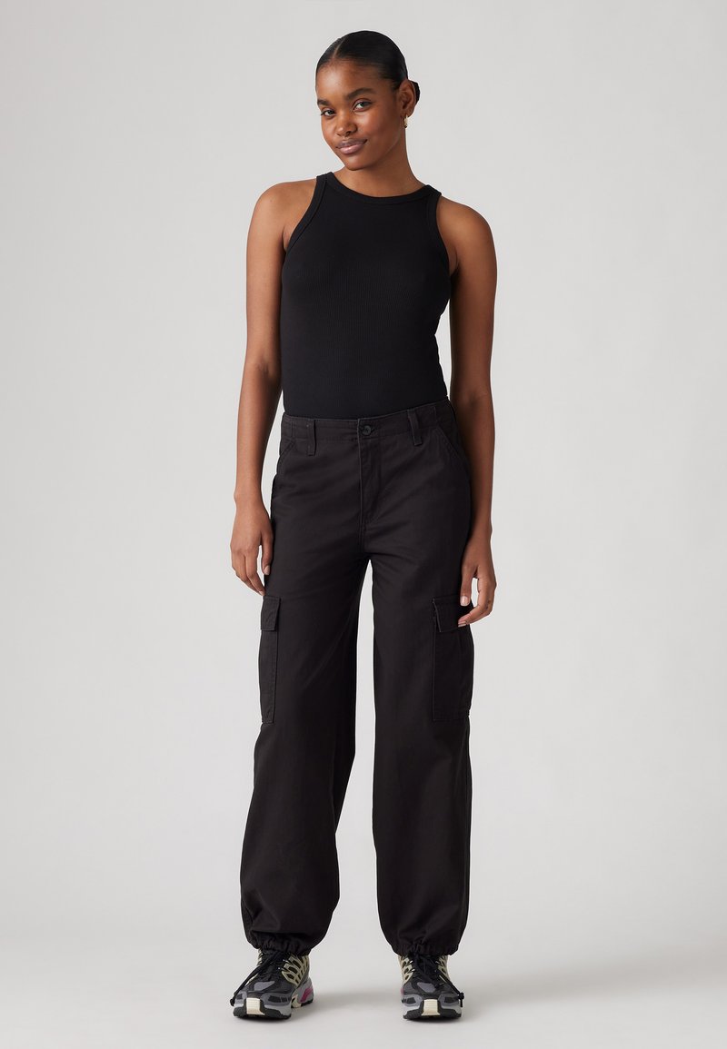 Débardeur noir à côtes associé à un pantalon cargo noir ample avec poches latérales, poignets froncés et chaussures de sport noires et grises.