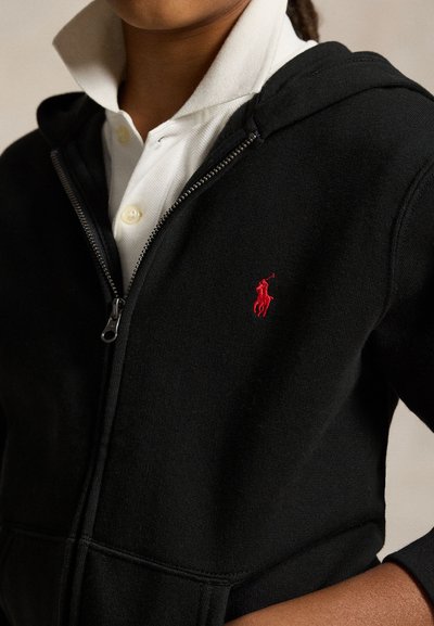 Person wearing a black zip-up hoodie with a red embroidered logo over a white collared shirt with the collar up.