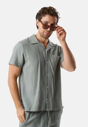 Mann trägt hellgraues, strukturiertes Kurzarm-Hemd mit Knöpfen und passende Hose, richtet seine Schildpatt-Sonnenbrille, vor schlichter Hintergrund.