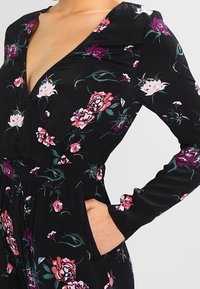 Czarny kombinezon w kwiaty z głębokim dekoltem w serek, długimi rękawami, kieszeniami i wszytą talią. Materiał ma gładką teksturę z różowymi i zielonymi wzorami.