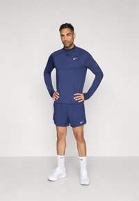 Μπλε ναυτικού χρώματος αθλητικό μπλουζάκι με μακριά μανίκια, ημικλείδωμα και λογότυπο· ταιριαστό σορτς· λευκά παπούτσια και κάλτσες· λείο ύφασμα, εφαρμοστή σχεδίαση.