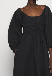 Svart klänning med fyrkantig halsringning, puffärmar och elastisk midja. Tillverkad av lätt material med en mjuk textur och flödande silhuett.