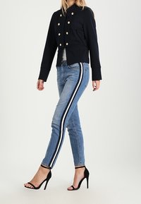 Marinblå kort jacka med guldfärgade knappar, i kombination med ljusblå jeans med vita sidostrimmor och svarta stiletthälar.