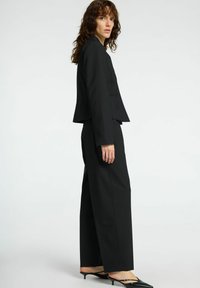 Černý elegantní oblek s krátkým blejzrem a širokými kalhotami. Hladká tkanina, strukturovaný design a minimalistický styl s špičatými černými podpatky.