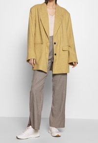 Gul rutigt oversize-blazer med stora slag, parat med en beige topp och bruna rutiga vida byxor. Vita sneakers fulländar outfitten.