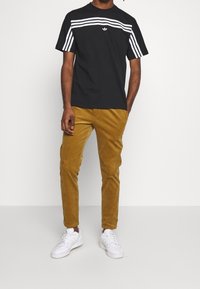 Camiseta negra con rayas horizontales blancas y logo en el pecho, combinada con pantalones de pana mostaza y zapatillas blancas.