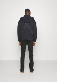 Svart huvtröja med ett strukturerat korsmönster på baksidan, i kombination med svarta byxor och svarta skor på en neutral bakgrund.