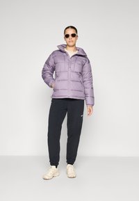 Lilla polstret jakke med glidelås og hette, kombinert med mørke joggebukser og beige sko. Modell som bruker solbriller. Enkel bakgrunn.