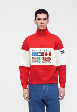 Ung mand iført en rød og hvid Tommy Jeans sweatshir med internationale flag og blå jeans står mod en ensfarvet lys baggrund.