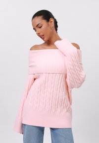 Sweater rosa de malha canelada com decote de ombro caído e punhos canelados, apresentando um ajuste solto e uma textura suave. Combinado com jeans azul claro.