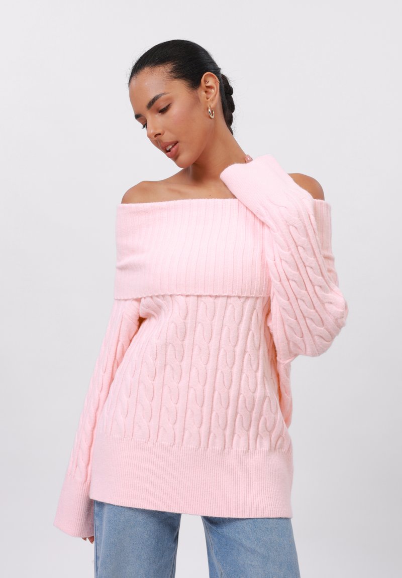 Sweater rosa de malha canelada com decote de ombro caído e punhos canelados, apresentando um ajuste solto e uma textura suave. Combinado com jeans azul claro.