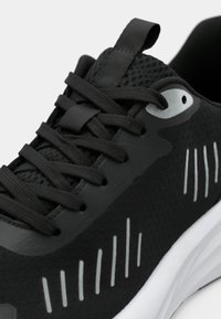 Gros plan d'une chaussure de sport noire avec des lacets noirs, une languette en mesh, des œillets argentés réfléchissants et des bandes réfléchissantes verticales sur le côté.