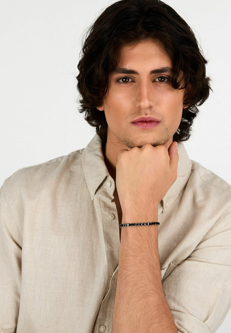 Bracciale di perle nere su un polso, composto da diverse perle circolari. Il portatore ha i capelli scuri e disordinati ed è vestito con una camicia di un beige chiaro.