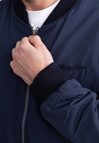 Personne fermant une veste bomber bleu marine avec col et poignets côtelés, portant un t-shirt blanc en dessous, main visible tenant la fermeture éclair.