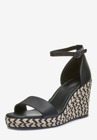 Sandalia de cuña de piel negra con tira en el tobillo, plataforma de yute trenzado con un patrón de espiga, textura suave y hebilla ajustable.