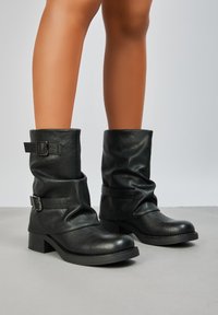 Bottines en cuir noir avec un design évasé, présentant des boucles sur les côtés, un talon épais et une finition texturée.