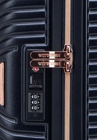 Gros plan sur le côté d'une valise rigide noire avec des tirettes de fermeture éclair en rose doré et une serrure à combinaison intégrée TSA007.