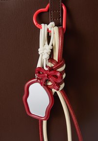 Bolso marrón con herrajes en forma de corazón rojos, correas texturizadas en crema y rojo, que presenta un lazo rojo y un pequeño accesorio de espejo.
