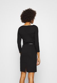 Robe noire avec un corsage ajusté, des manches trois-quarts et une fente dans le dos. Fabriquée dans un tissu lisse avec un léger éclat et une taille ceinturée.