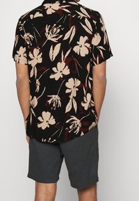 Mustavalkoinen, lyhythihainen paita, jossa on beigen värinen kukkakuvio, kaarevat helmareunat ja rento istuvuus, yhdistetty tummanharmaisiin shortseihin.