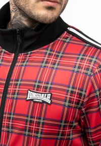 Crvena karirana jakna na zip s crnim detaljima, koja ima uspravan ovratnik i Lonsdale logo zakrpu. Tkanina je glatka s sjajnim finišom.