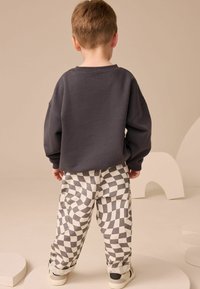 Dziecko stoi plecami do kamery, ubrane w ciemny, za duży sweatshirt oraz czarno-białe dżinsy w kratę z podwiniętymi nogawkami i trampkami.