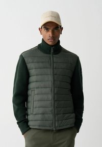 Paminkštintas žalias liemenė su užtrauktuku, su riešo mezgimo rankovėmis ir aukšta apykakle. Modelis dėvi kreminės spalvos kepurę su siuvinėtu tekstu.