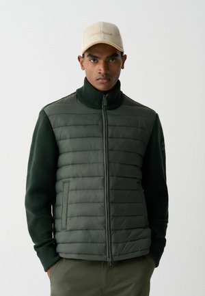Vestă verde căptușită cu închidere prin fermoar, mâneci din tricot cu nervuri și guler înalt. Modelul poartă o șapcă bej cu text brodat.