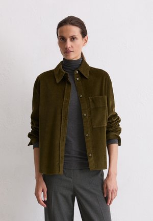 Olijfgroene ribfluwelen shirt met een knoopsluiting aan de voorkant, puntige kraag en borstzak, gelaagd over een donkergrijze coltrui. Gestructureerde stof.