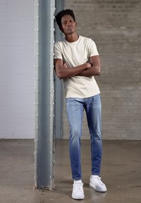 T-shirt beige in cotone abbinata a jeans di denim blu chiaro e sneakers bianche, appoggiato contro un pilastro metallico in un ambiente minimalista.